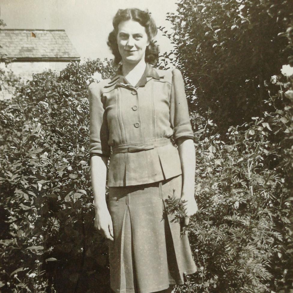 A black and white photo of a woman smiling at the camera in front of bushes 