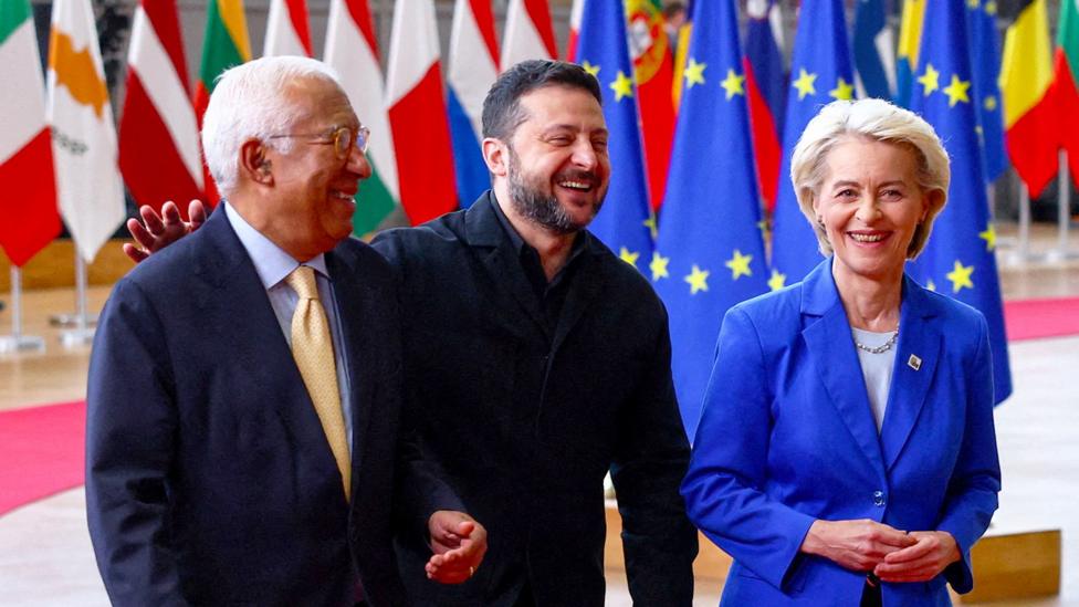 António Costa, Volodymyr Zelensky and Ursula Von der Leyen laughing as they walk, with many country flags hanging behind them.