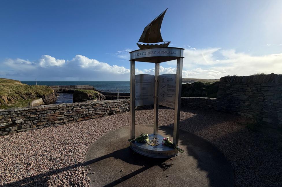 The memorial is a tall metal structure with a plinth and three narrow legs holding an upper section with the sculpture of a fishing boat with a sail. The memorial is in a gravel-covered area at the harbour at Port of Ness.