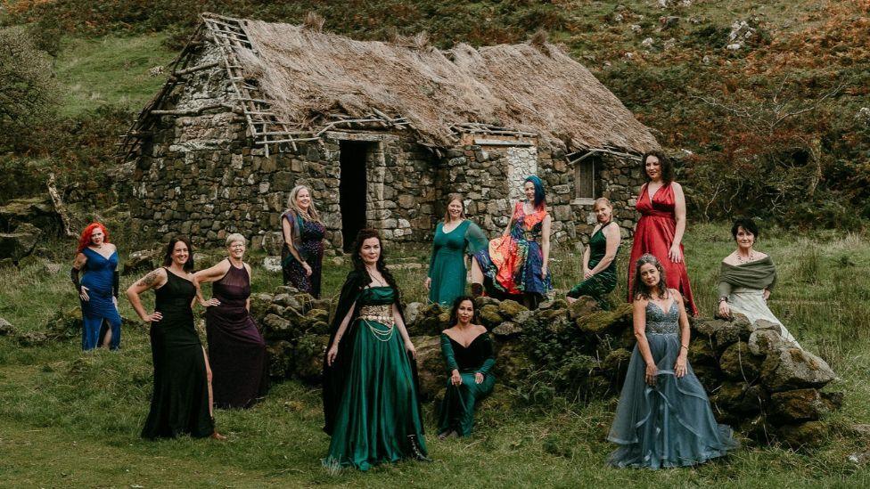 A group of 11 women in colourful gowns pose around a low stone wall in front of a traditional stone cottage with a thatched roof. The cottage sits in a grassy rural landscape surrounded by trees and hillside vegetation, while the people are spaced across the scene in various standing and seated positions.