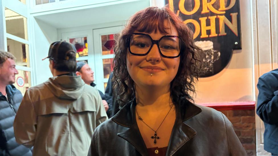 Jade is a young woman with a bob wearing a leather jacket and glasses. She smiles to the camera in a bar surrounded by other young people.