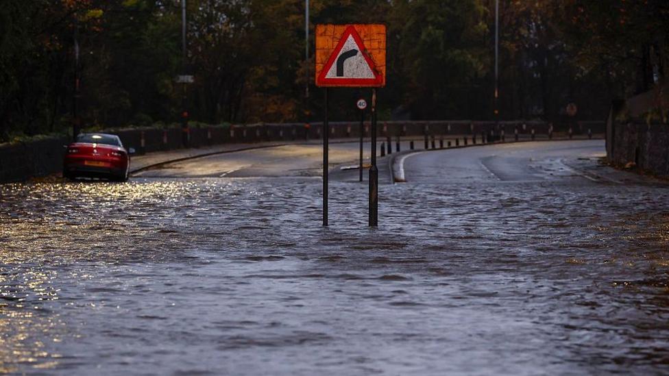 A flooded road with a right-hand bend sign in the middle of it and a car stuck in the water.