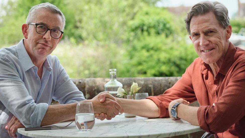 Steve Coogan, left, and Rob Brydon, right, are sat across the table from each other and shaking hands. They are both smiling at the camera.