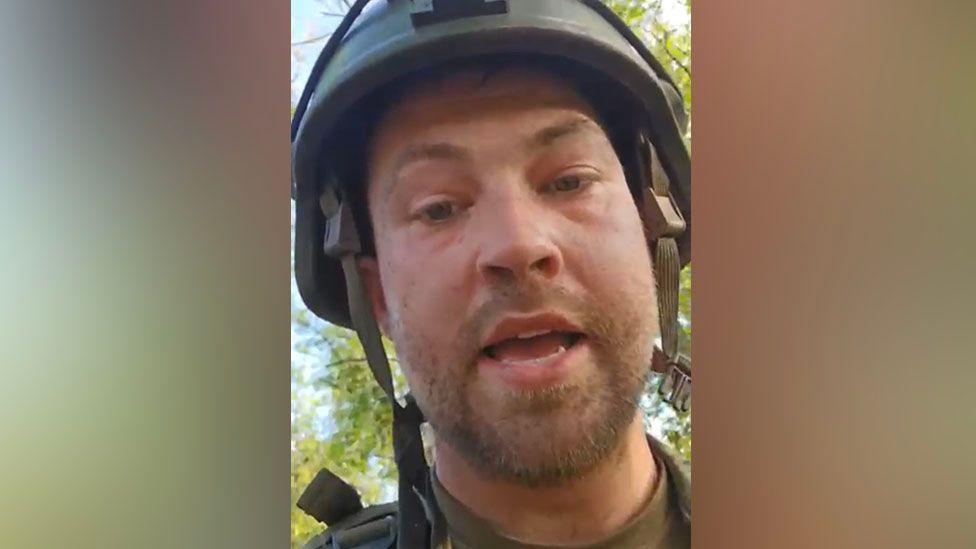 Натан Стоббарт одет в хаки военную форму и на нём бронешлем с незастёгнутым ремешком под подбородком. У него щетина на бороде и усах, мешки под глазами, он выглядит уставшим. Рот открыт, словно он что-то говорит. Позади него зелёные лиственные деревья и бледно-голубое небо.