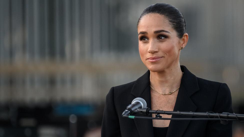 Meghan Markle stands behind a microphone wearing a simple charcoal jacket and top, a gold necklace hangs across her chest. Her dark hair is pulled back in a bun.