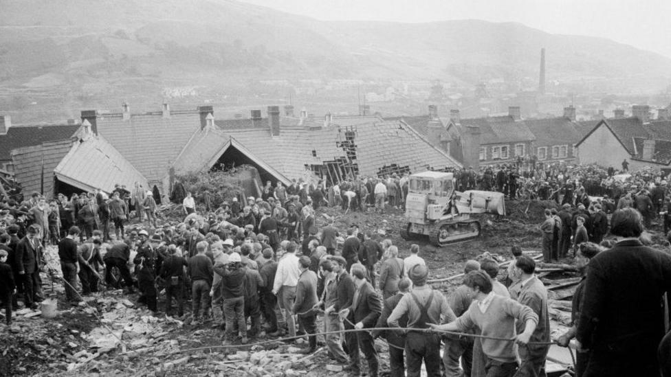 Black and white picture shows the mud and devastation caused at Pantglas Junior School on 21st October 1966. A row of men pull a rope, with the submerged school with a collapsed roof in the background. Crowds are gathered around the scene.