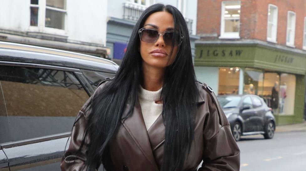 Katie Price wearing a brown leather coat, cream jumper and dark sunglasses standing near a road.