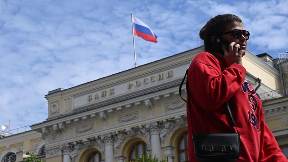 Qumal rang koʻylak va quyoshdan saqlanuvchi koʻzoynak taqqan, oʻng yelkasida kichik sumkasi boʻlgan ayol Rossiya Federatsiyasi Markaziy banki binosining tashqi koʻrinishi oldida telefon bilan gapirmoqda, binoning ustida Rossiya bayrogʻi koʻrinib turibdi