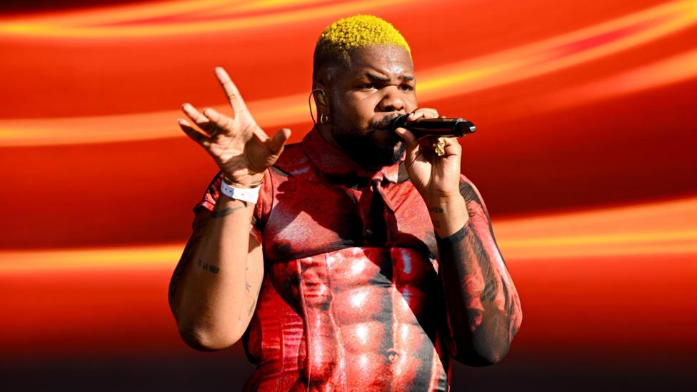 MNEK sahnada chiqish paytida mikrofonga qo'shiq aytmoqda. U muskulli tana rasmi bosilgan qizil ko'ylak kiygan va sochlari sariq.