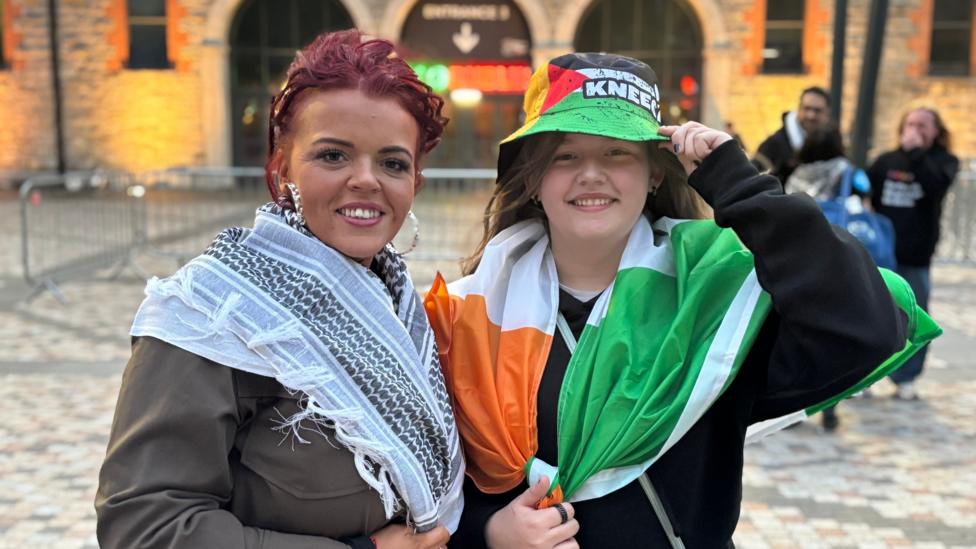 Nicola McCollough, who has red hair tied back and wears silver-coloured hoop earrings, a keffiyeh scarf and a brown jacket. Her daughter Lily Rose stands to her left wearing a black hoodie, a bucket hat in the colours of the Palestine flag and with the Kneecap logo on it and an Irish tricolour around her shoulders.