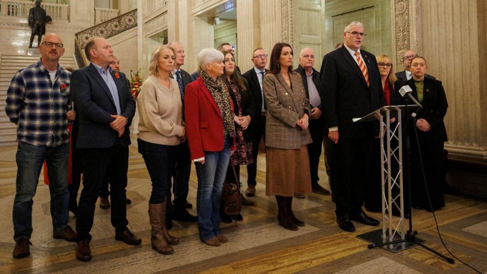 Stormontdagi Buyuk zalda bir nechta DUP siyosatchilari turibdi. Geyvin Robinson mikrofon oldida turibdi. U qora kostyum va qizil-sariq chiziqli galstuk kiygan.