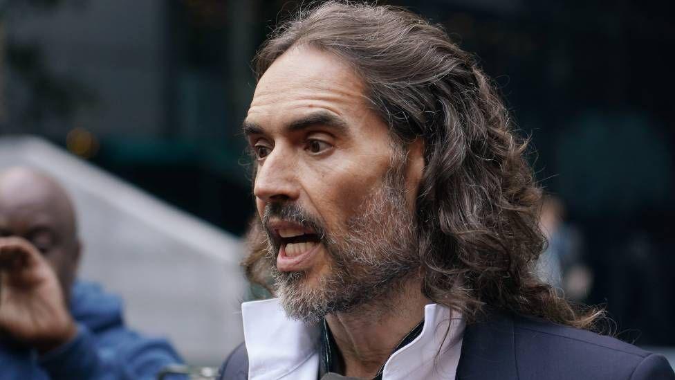 Rassel Brend 2026-yil 28-oktyabrda Angliyaning London şahridagi Sautuark toj sudida zörlaş va jinsiy zöravonlik ayblovlari bilan körilayotgan jarayonidan oldin sudga kelmoqda
