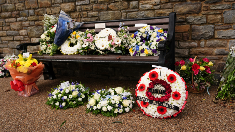 A number of flowers and wreaths have been placed beside and on a bench next to a stone wall.