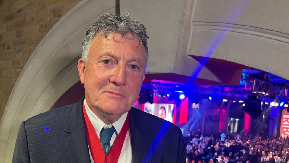 Riba president Chris Williamson wearing a red ribboned medallion and standing in the foreground as an awards ceremony goes on behind him.