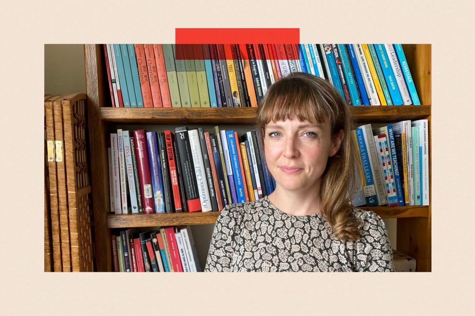 Lucy Crehan stands against a bookcase. She has long brown hair and is wearing a patterned top