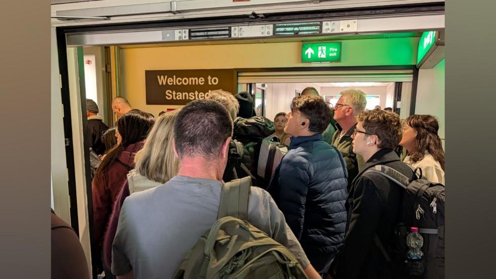 A number of people queuing in a corridor. There is a sign that reads “Welcome to Stansted” hanging on the wall.