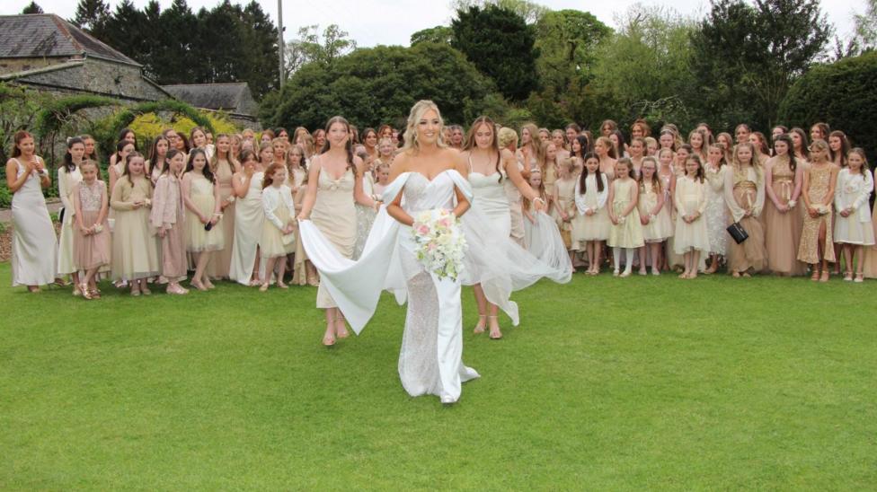 A bride, with blonde hair, a white dress with an off the shoulder detail carrying a bouquet, smiles as two young bridesmaids help her with her train and veil as she walks towards the camera. Behind them is a large group of women, including young girls in the front row, wearing a mixture of champagne, cream, white and tan dresses. The crowd, including the bride and the two bridesmaid, are standing in the middle of a large garden, with tall trees and a leafy tunnel to the left.