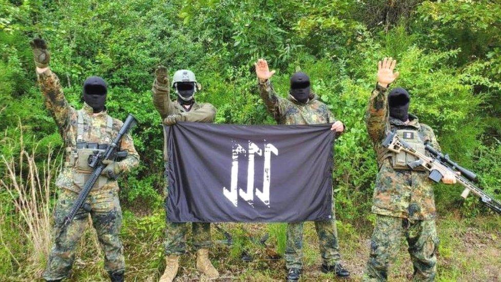 Мужчины в армейской форме и масках держат неонацистские флаги в сельской местности