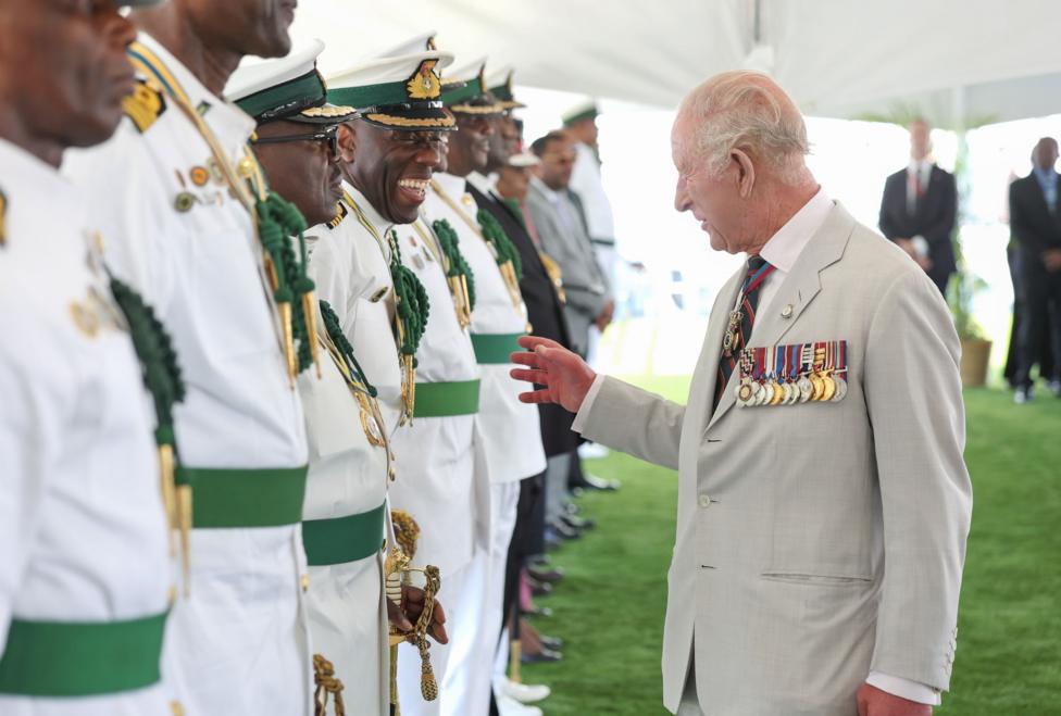 King Charles, wearing a light beige suit with military medals displayed speaks to veterans wearing white uniforms highlighted with green