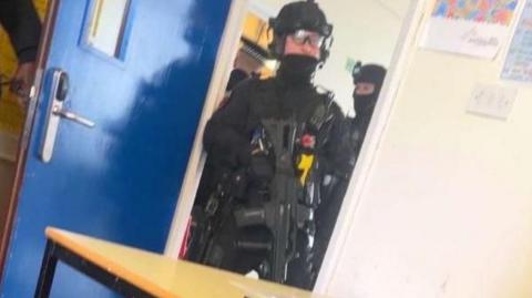 Вооружённый полицейский стоит у двери в класс. Он одет во всё чёрное, в шлеме и очках. Он держит большой чёрный пистолет, направленный в пол