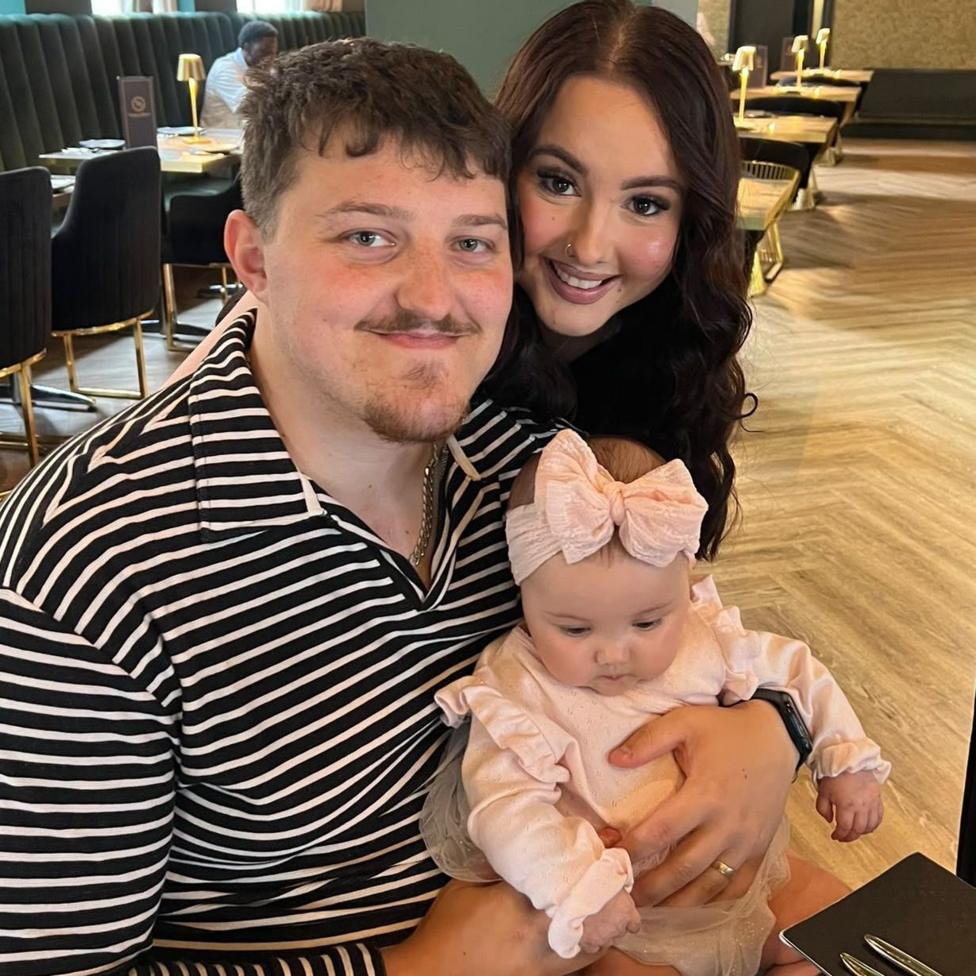 Sophie and Ryli Price are sitting in a restaurant with their baby daughter Florence. Ryli has a black and white horizontal striped shirt on and has short light brown hair with a goatee. Sophie is to his left and has long, dark wavy hair and a nose piercing. Florence is on her dad's lap and is wearing a light pink jumper with frilly cuffs and a matching bow on her head.
