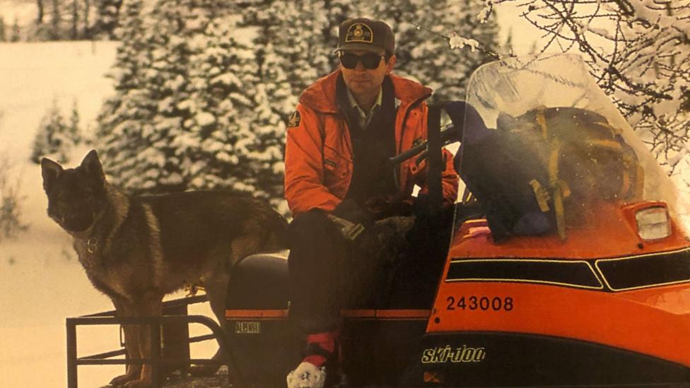 An image showing Scott Ward sitting on a snowmobile with a German Shepherd beside him. He is wearing an orange coat with a park warden hat and dark pants. Behind him are snowy trees.