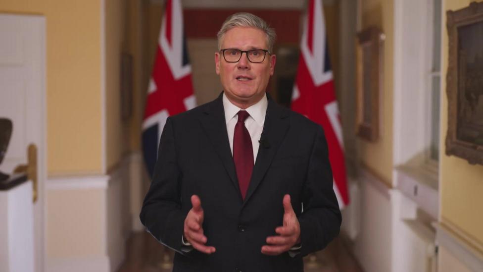 Kir Starmer Buyuk Britaniya bayroqlari fonida turib kameraga qarab sözlaşmoqda