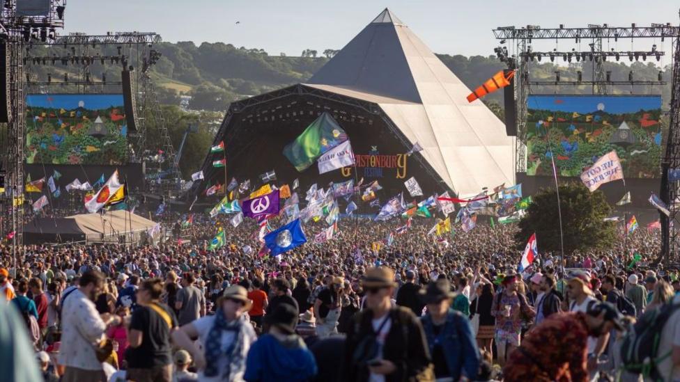 A large crowd in front of the Pyramid Stage at Glastonbury Festival. Lots of people are holding up large flags and waving them as the sun sets over the stage.