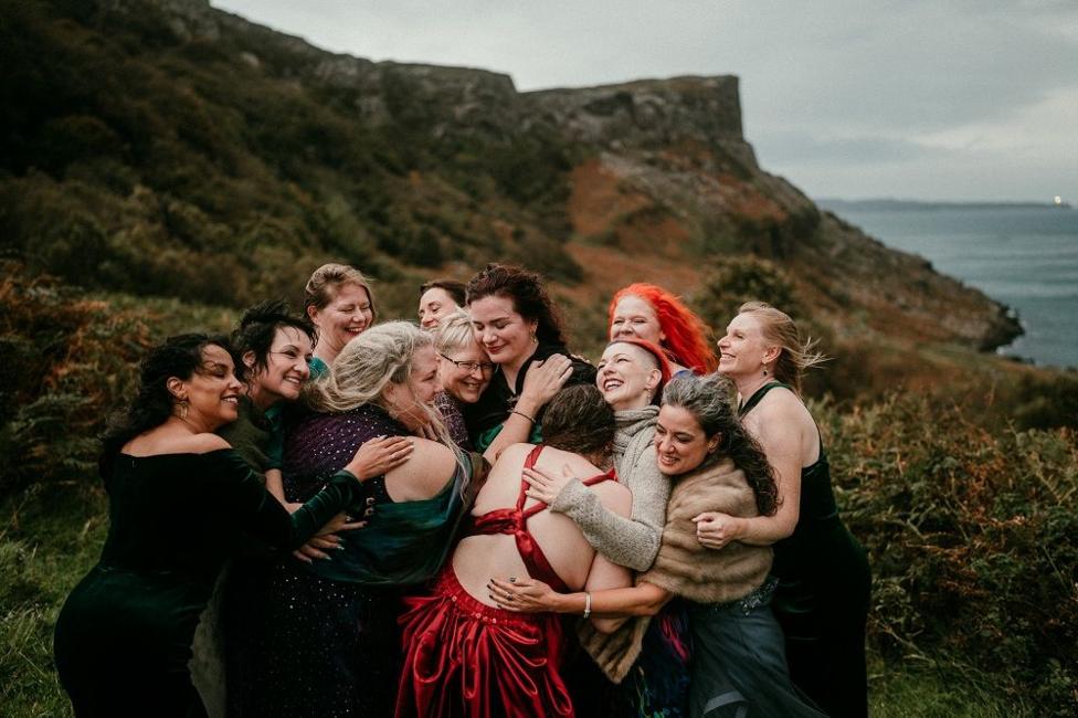 A group of 10 women stand in a close embrace on a grassy hillside overlooking the sea. Several wear formal dresses in dark and red colours. Behind the group, rugged coastal cliffs rise under an overcast sky.