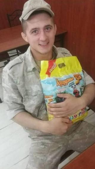 Artyom is in army camos and a cap sitting down. He is holding yellow packaging 