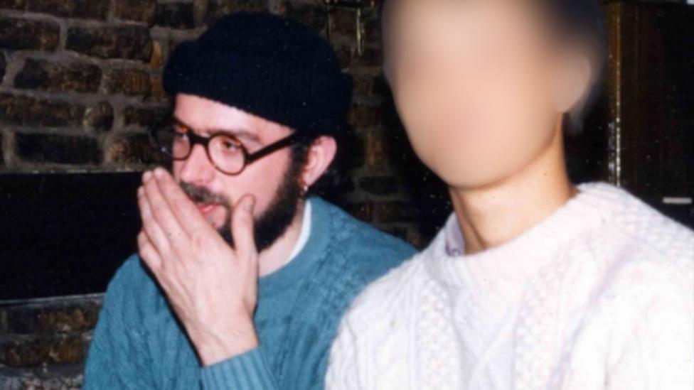 An image of Andrew Coles in a blue knitted jumper, round-rimmed spectacles, a black beanie hat and beard. A woman in the foreground has been blurred out to protect her identity.