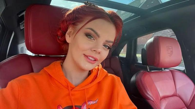 A woman with red hair looks at the camera. She is wearing an orange hoodie and is sitting inside a car.