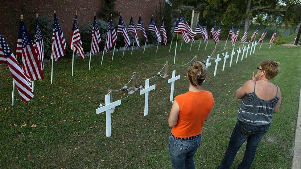 A line of white crosses are stood in grass alongside American flags while two women's backs are shown as they look at the crosses
