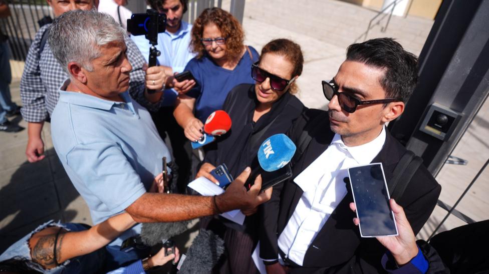 Alexandros Alexandrou, who is wearing a suit and black sunglasses, speaking to a crowd of reporters outside court.