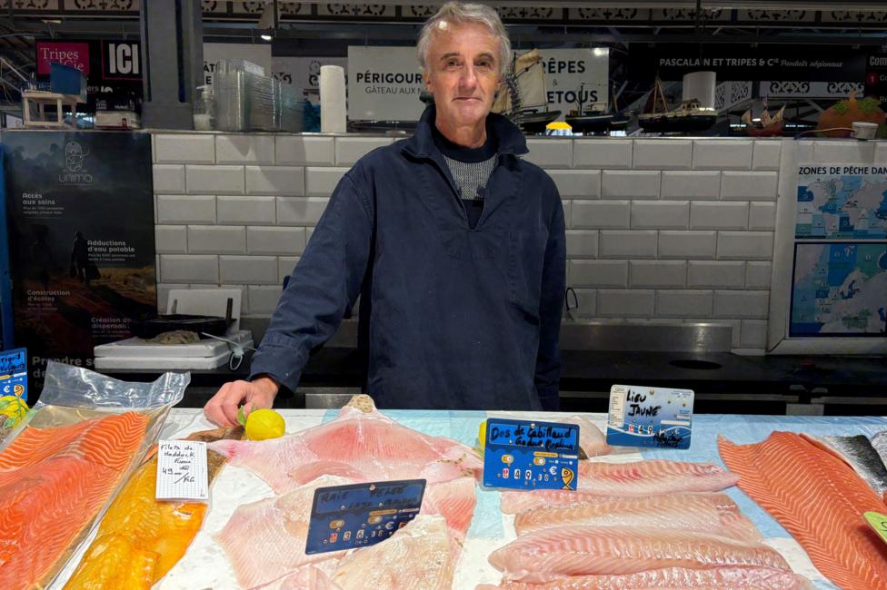 Jean Roger Faucher, a man in his sixties, stands behind his fishmonger's stall. Various cuts of fish are in front of him and he wears a navy jacket.