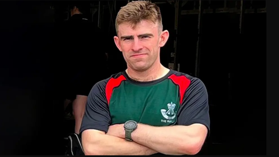 A young man with blonde hair. He stands with his arms crossed. He is wearing a t-shirt which says 'the rifles' and he wears a black watch. He looks strong.