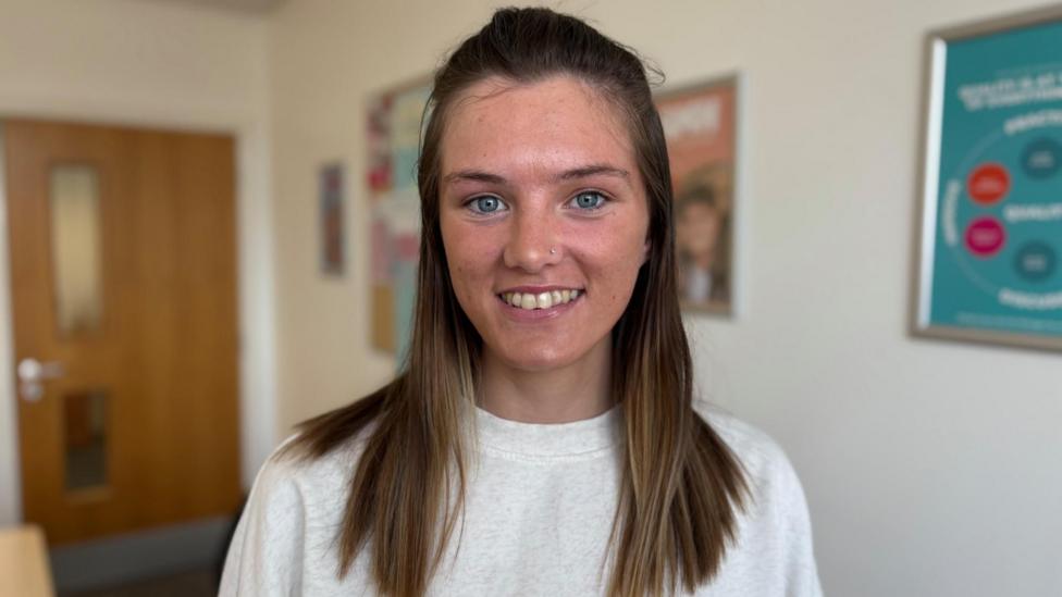 Close-up portrait of Neala with shoulder-length brown hair wearing a light-coloured sweatshirt, standing indoors. A wooden door and framed posters are visible in the softly blurred background.