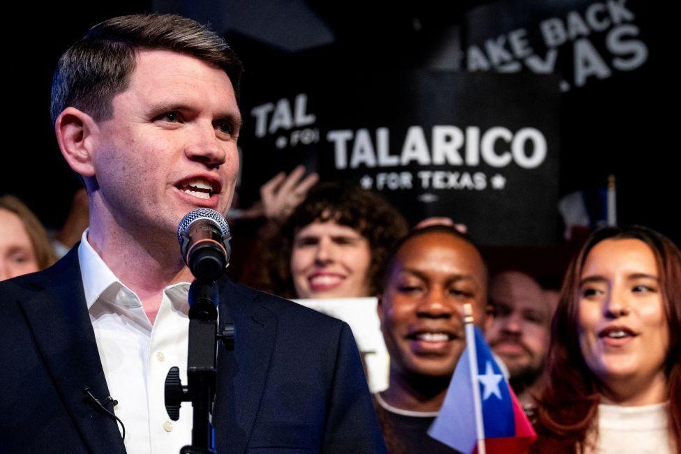 James Talarico, wearing a sports coat and collared shirt with the top button undone, speaks in front of a crowd