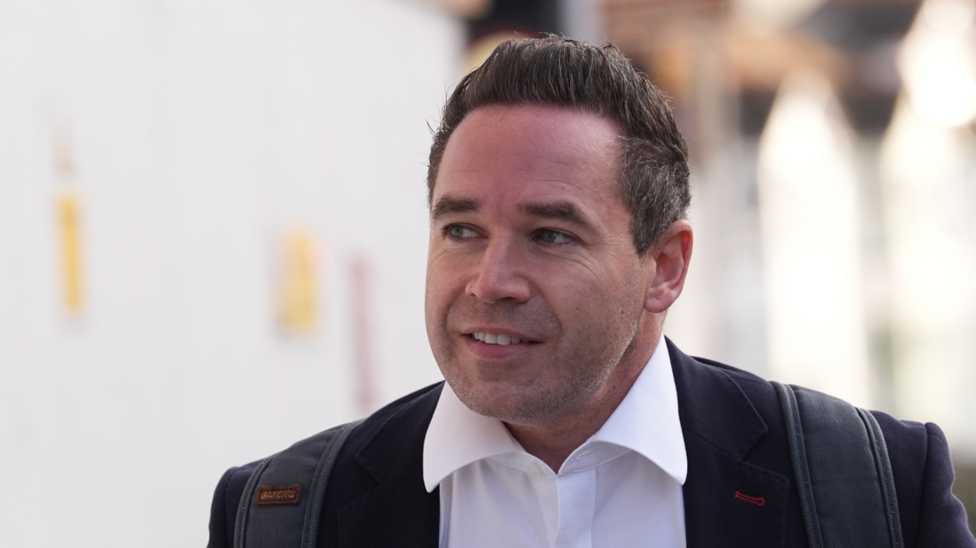 A man with short black hair wearing a suit jacket and white shirt. He is looking away from the camera.