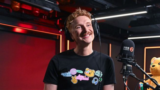 BBC Radio 2 boshlovchisi Owain Wyn Evans. Quloqchinlar va qora futbolka kiyib, mikrofon oldida tabassum qilmoqda