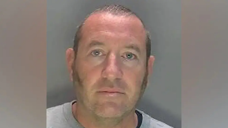David Carrick photographed in police custody. He is wearing a grey sweatshirt and has sideburns and a near-shaven head.