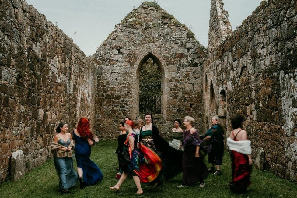 A group of women in colourful dresses move through the roofless stone interior of a historic ruin. One person stands near the centre in a green dress and black cloak, while others walk around her, creating a sense of motion. Tall stone walls and a pointed arched window frame the scene, with grass covering the ground inside the building.