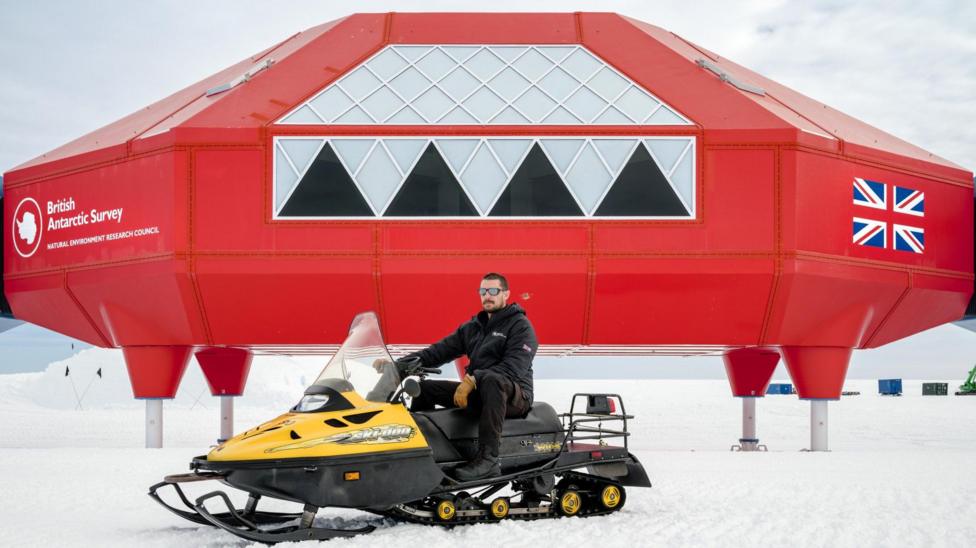 Den Makkenzi Antarktidagi Xolli VI tadqiqot stansiyasi oldida qor motosiqlida (snowmobile) o‘tirgan holda