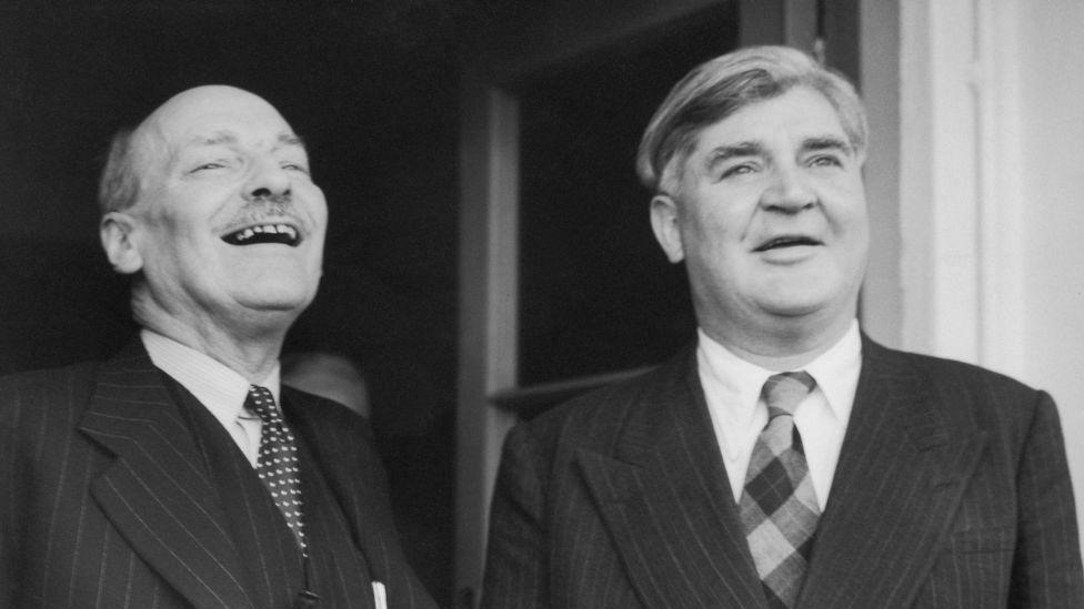 Clement Attlee (chapda) va Aneurin Bevan (o'ngda). Clement boshini orqaga tashlab kulmoqda, Bevan esa to'g'riga qarab tabassum qilmoqda. Bu ikki erkakning cho'zilgan kostyum, ko'ylak va galstuk kiygan, fonda ochiq eshigi bor qora-oq rasmi.