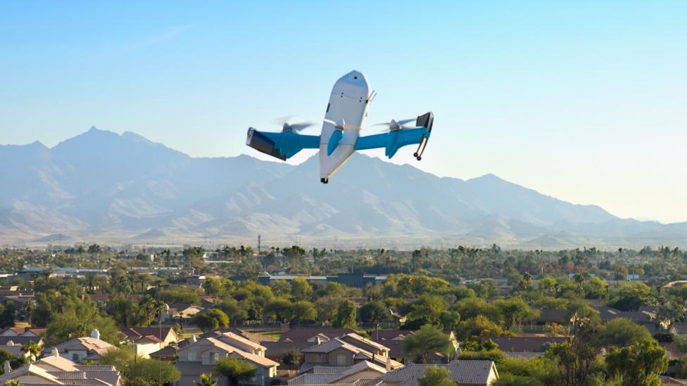 An Amazon drone flies over a US city. In the background are mountains and a blue sky.