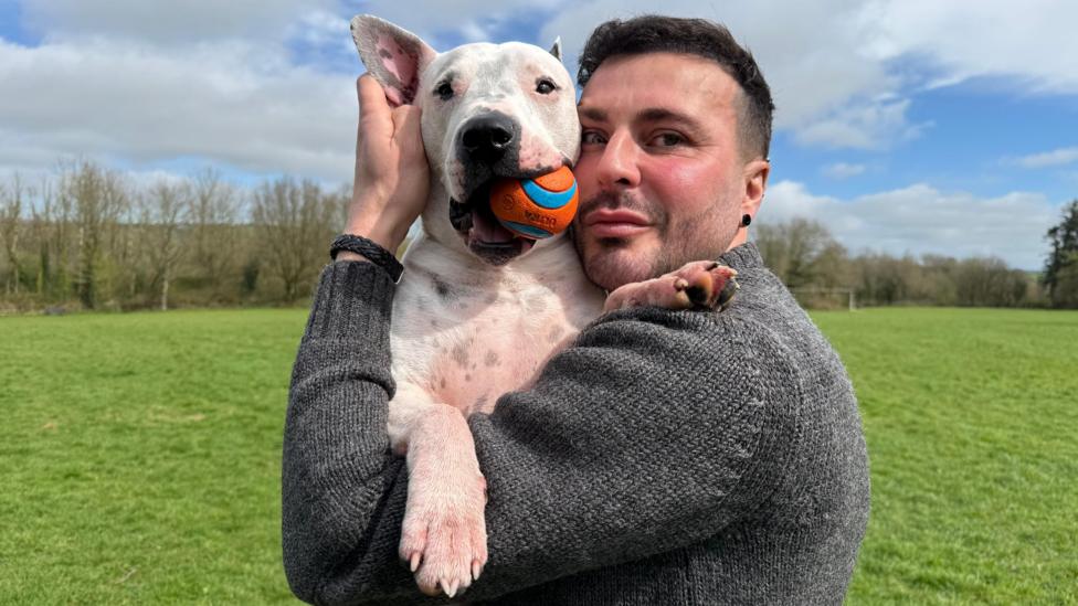 James is standing in a grass field holding his dog which has a ball in his mouth. James wears a grey jumper and looks at the camera.