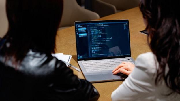 The image shows two people from behind as they sit at a table looking at code on a laptop.