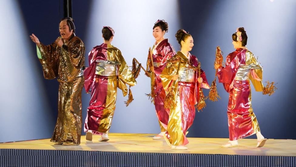 A man and four women in shiny pink-gold traditional-style robes stand under bright stage lighting, holding decorative gold props - part of a performance at the Asian Games 2026 closing ceremony