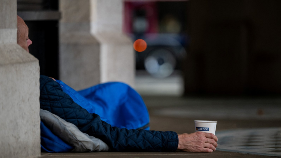 A homeless person lies in a London doorway in a blue sleeping bag. Part of the man's face is visible around the corner of a building and he is holding a paper cup.