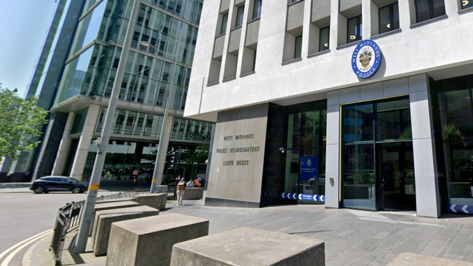 The outside of a police headquarters building with an oval badge above a set of glass doors identifying it as the West Midlands Police force. There is a road surrounding the building and other tall buildings nearby.